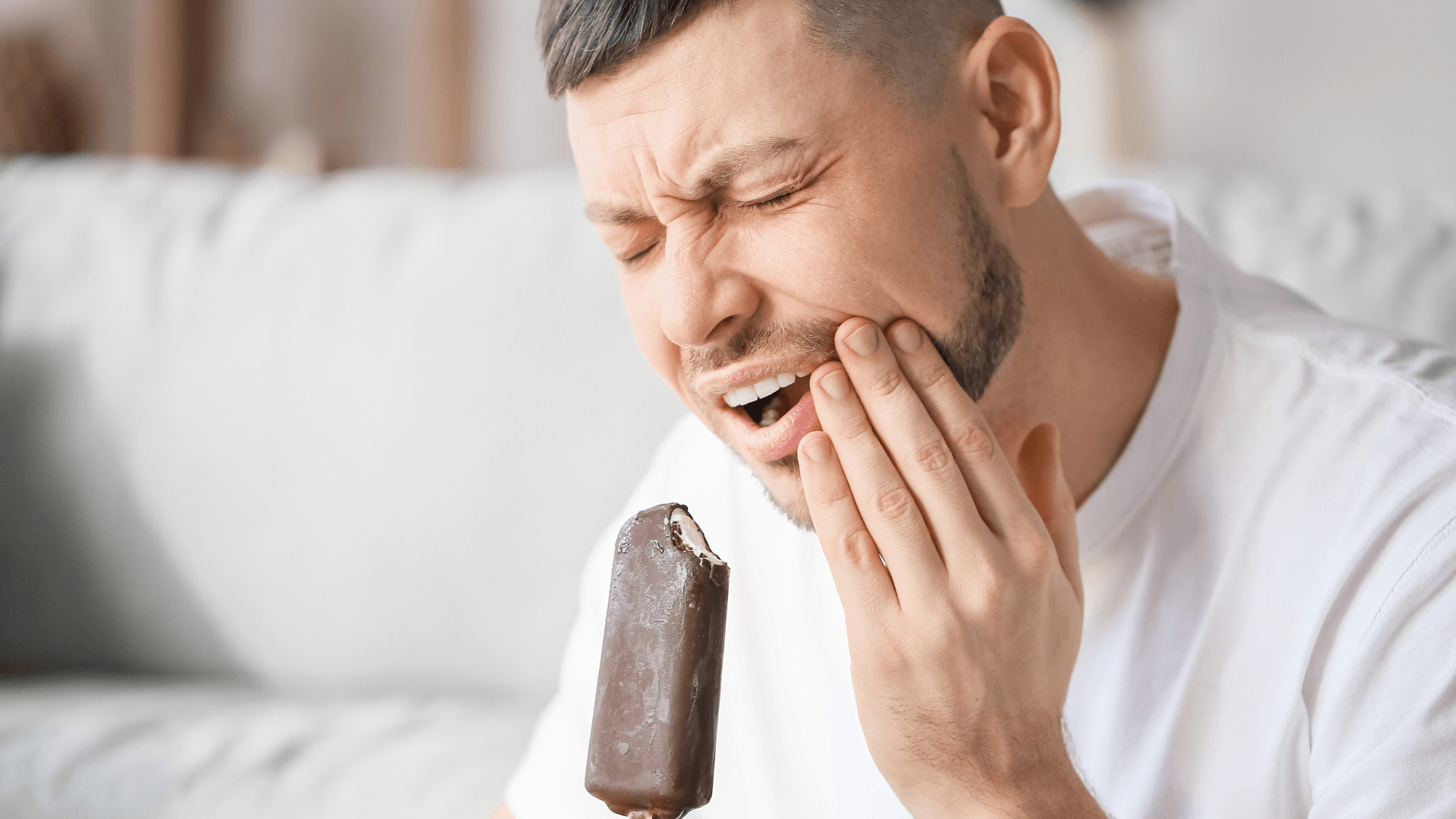 Primer plano de una persona llevándose la mano a la mejilla con expresión de dolor agudo al intentar comer un helado, sufriendo un pinchazo por sensibilidad dental al frío.